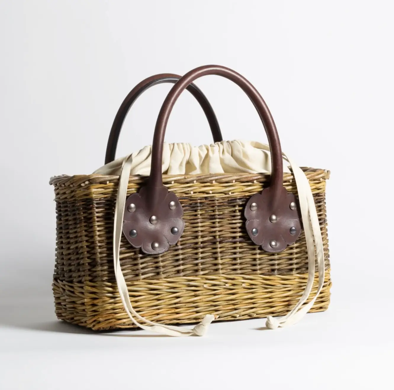 A structured wicker handbag with a rectangular silhouette, featuring woven straw construction in natural tones and sturdy brown leather top handles secured with riveted floral-shaped patches. The interior is lined with a soft, gathered cream fabric, with long drawstring ties draping over the sides. The design blends rustic craftsmanship with refined detailing, giving it a polished yet artisanal feel