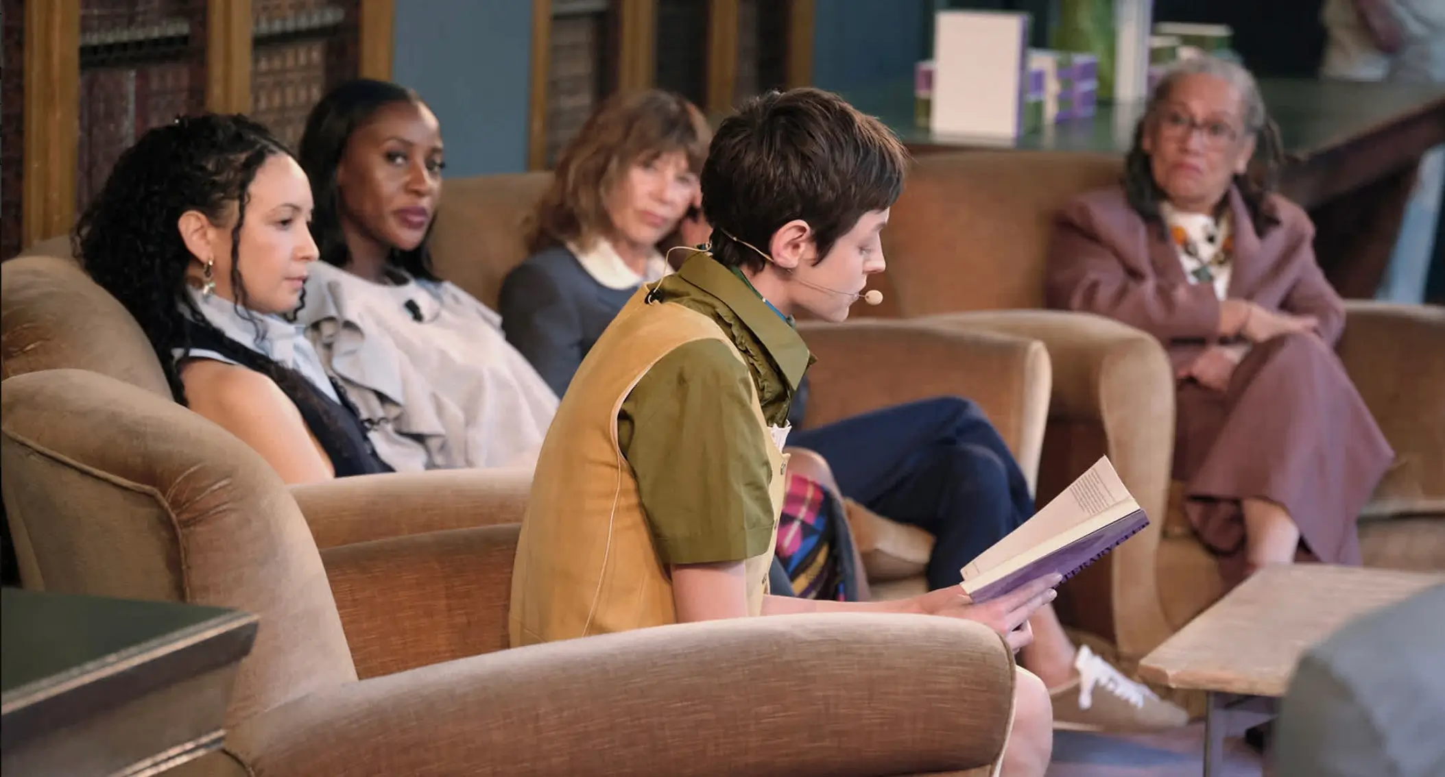 A small, intimate panel setting with five women seated in plush armchairs, arranged in a relaxed semicircle within a warm, library-like space. In the foreground, a speaker wearing a headset microphone reads from a book, leaning slightly forward with focused attention. Behind her, the other participants—dressed in understated, tailored looks—listen quietly, their expressions attentive and reflective. The setting, with wooden shelving and soft lighting, feels conversational and intellectual, blending the tone of a literary reading with a curated cultural discussion