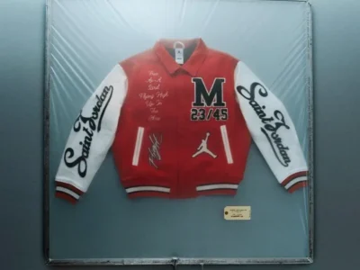 Red SAINT Mxxxxxx x Jordan Brand varsity jacket encased in a sealed display frame, featuring white sleeves with “Saint” script lettering, a bold “M 23/45” chest patch, handwritten-style “Free As A Bird Flying High Up In The Skies” text, Jumpman logo detailing, and striped ribbed trims, presented against a muted grey backdrop