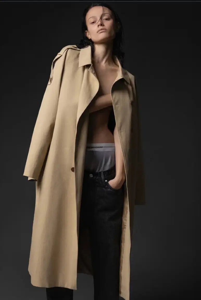 Studio portrait of a model standing against a dark backdrop, wearing an open beige trench coat draped loosely over the shoulders, revealing a bare torso and low-rise dark jeans with a visible waistband. The model’s hair appears wet and slicked back, with a relaxed, slightly tilted pose and one hand in the pocket, conveying a minimalist, high-fashion editorial mood