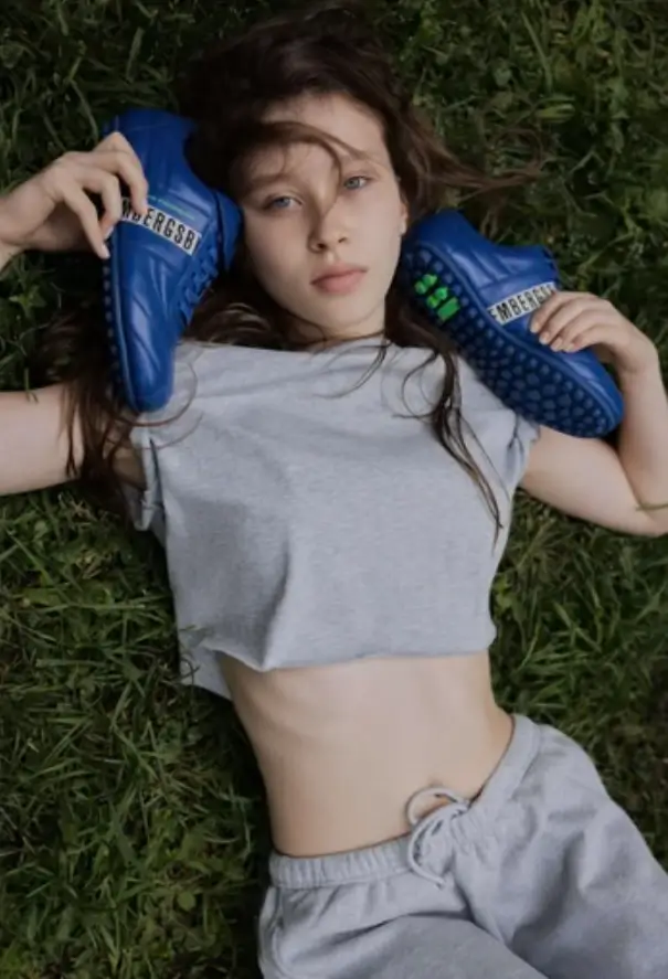Overhead shot of a person lying on green grass, wearing a cropped gray T-shirt and matching sweatpants. They hold a pair of bright blue athletic shoes on either side of their head, with one resting near the ear, creating a relaxed, editorial-style composition