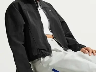 Woman seated wearing Nike Airreverent black cropped zip-up jacket over a white top, paired with light gray wide-leg pants featuring a bold blue side stripe. Minimalist techwear styling from the Airreverent collection