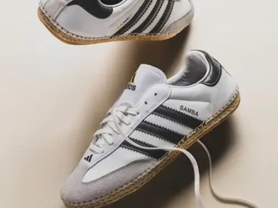 angled product shot of adidas x CLOT Samba shoe in white with black Three Stripes, featuring suede toe caps, gold “Samba” branding, espadrille-style woven soles, and loose laces against a neutral backdrop