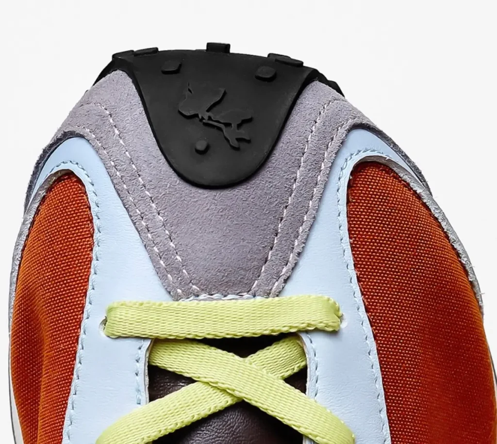 Close-up of shoe upper showing orange mesh panels, light blue leather overlays, grey suede center panel with black rubber heel tab, and neon yellow laces crossing over the tongue