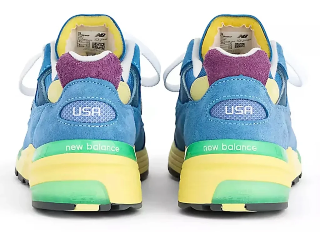 A rear view of the New Balance 992 “Sea Stone” Made in USA shoe, showing both heels side by side. The design features blue suede uppers with purple heel tabs, yellow interior lining, and “USA” branding on the back. The layered midsole combines pale yellow and green tones, with “new balance” text along the heel, highlighting the shoe’s multi-color construction against a clean white background