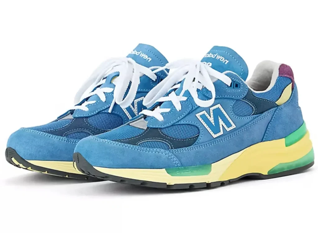 A three-quarter view of the New Balance 992 “Sea Stone” Made in USA shoe angled slightly outward. The design features a blue-green suede and mesh upper with white laces and the signature “N” logo, paired with a layered midsole in pale yellow and green with subtle purple accents at the heel. The composition highlights the silhouette’s balance of classic running form and modern color detailing against a clean white background