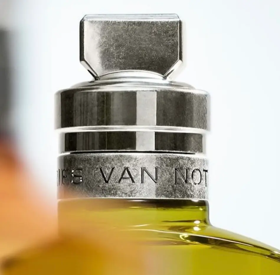 Extreme close-up detail of the Dries Van Noten Crazy Basil Eau de Parfum bottle cap. A heavy, faceted matte silver metal cap with geometric edges sits atop a polished metallic collar engraved with “DRIES VAN NOTEN.” The luxurious brushed-metal texture contrasts beautifully with the vibrant olive-green fragrance liquid visible below, highlighting the brand’s signature refined craftsmanship and minimalist luxury aesthetic