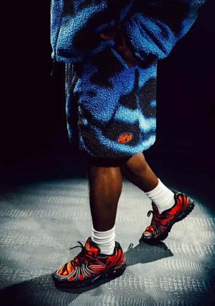 Person in blue-black camo fleece jacket and shorts wearing bold red and black New Balance shoe with white socks