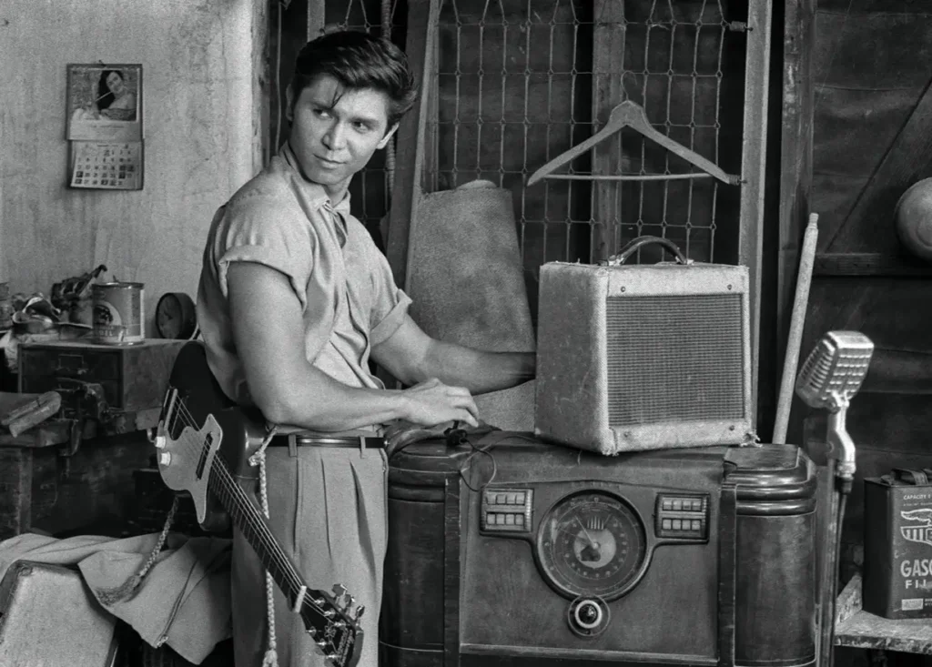 A young man stands beside a vintage radio and amplifier with a guitar nearby, adjusting equipment in a black-and-white interior scene