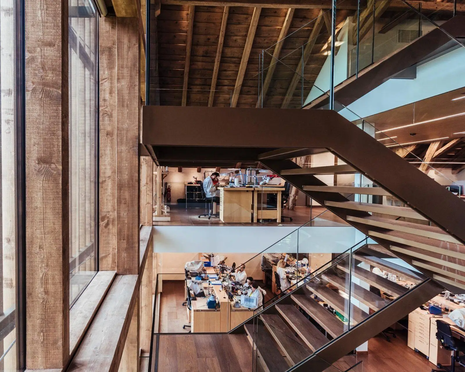 Modern watchmaking atelier with open-plan wooden interior, featuring artisans at workstations across two levels connected by a sleek metal staircase, combining traditional craftsmanship with contemporary architectural design