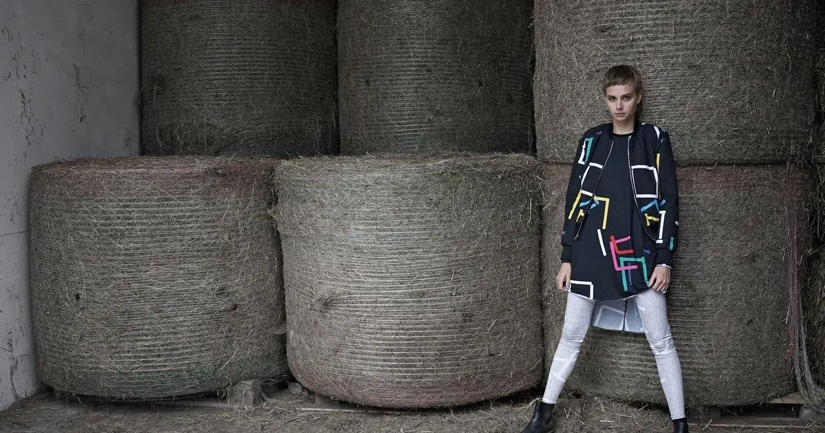 Model standing in front of stacked hay bales wearing a graphic patterned jacket, long tunic top, and light pants, set in a rustic industrial barn setting