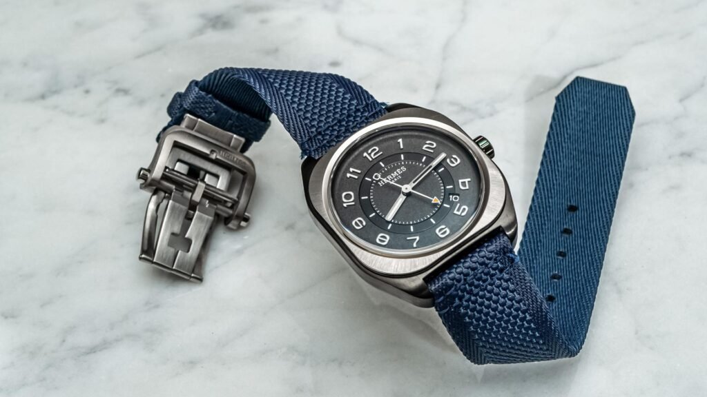 Hermès H08 watch with a brushed steel cushion-shaped case and grey dial, paired with a textured navy fabric strap, displayed on a marble surface alongside its detached metal folding clasp
