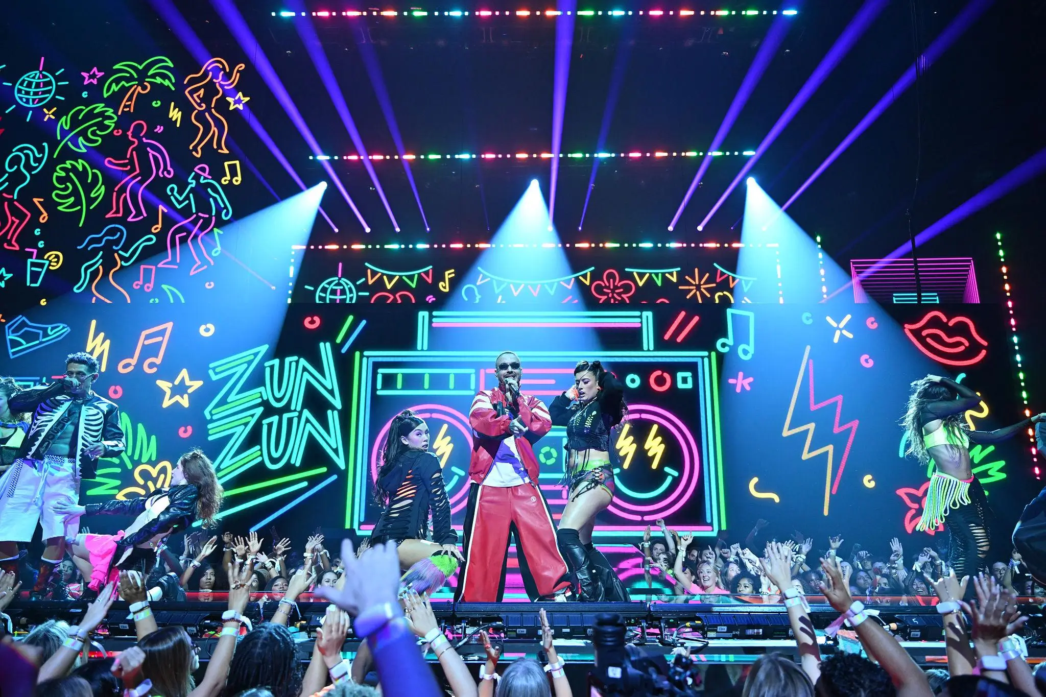 A high-energy concert scene with a male performer in a red outfit at center stage, flanked by dancers in bold, neon-accented costumes. Behind them, a massive LED backdrop displays colorful graffiti-style graphics, including a large boombox and the words “ZUN ZUN.” Bright stage lights beam outward in vivid purples and blues, while a packed crowd in the foreground raises their hands, capturing the electrifying atmosphere of a live music performance