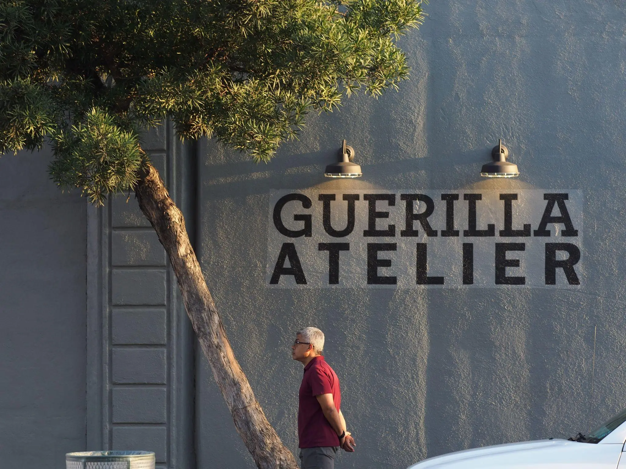 Man walking past a gray exterior wall with large “GUERILLA ATELIER” lettering, framed by a leaning tree and overhead industrial lights, with a parked car partially view in the foreground