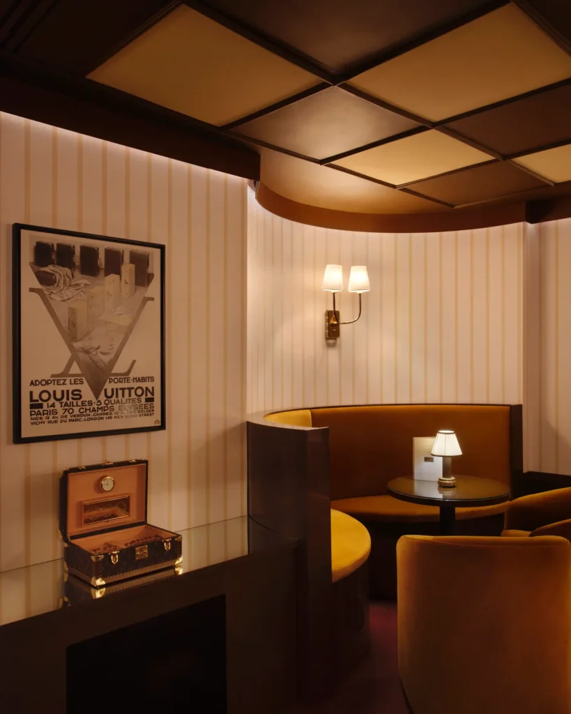 An intimate lounge-style Louis Vuitton interior featuring a curved velvet banquette in warm amber tones set against striped walls and soft ambient lighting. A small round table with a lit lamp sits at the center, while a vintage-inspired Louis Vuitton poster and an open monogram trunk display add heritage detail. The coffered ceiling and brass wall sconces enhance the refined, boutique atmosphere, evoking a quiet, private salon within a haute retail space