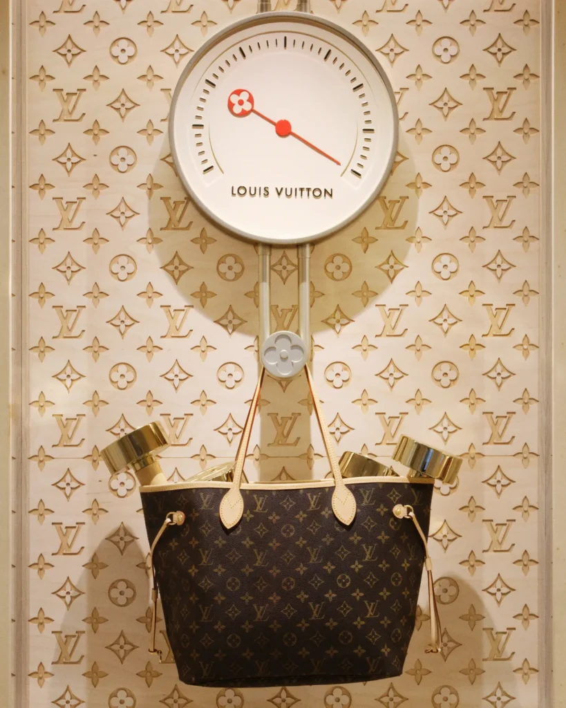 A conceptual Louis Vuitton display featuring a classic monogram tote suspended beneath a stylized wall-mounted scale or clock with red hands and LV detailing. The bag, filled with gold-toned weights, hangs against a backdrop of repeating monogram motifs, blending fitness-inspired imagery with luxury iconography in a way, design-forward retail installation