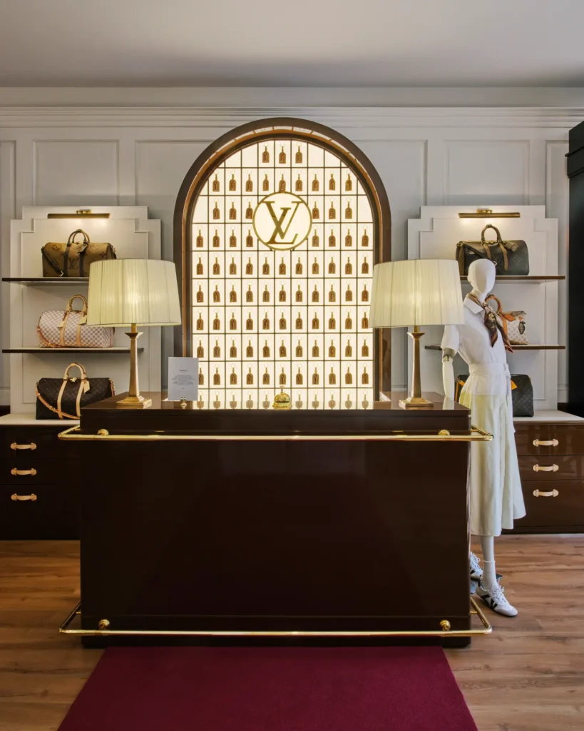 A refined Louis Vuitton boutique interior featuring a polished dark wood service counter centered beneath a glowing arched display wall adorned with gold accents and the LV monogram. Symmetrical shelving on both sides showcases classic monogram handbags, while two cream-shaded lamps frame the scene. To the right, a minimalist mannequin styled in a white outfit with a silk scarf stands near the display, set against paneled walls and warm wood flooring, evoking a quiet, high-end retail atmosphere