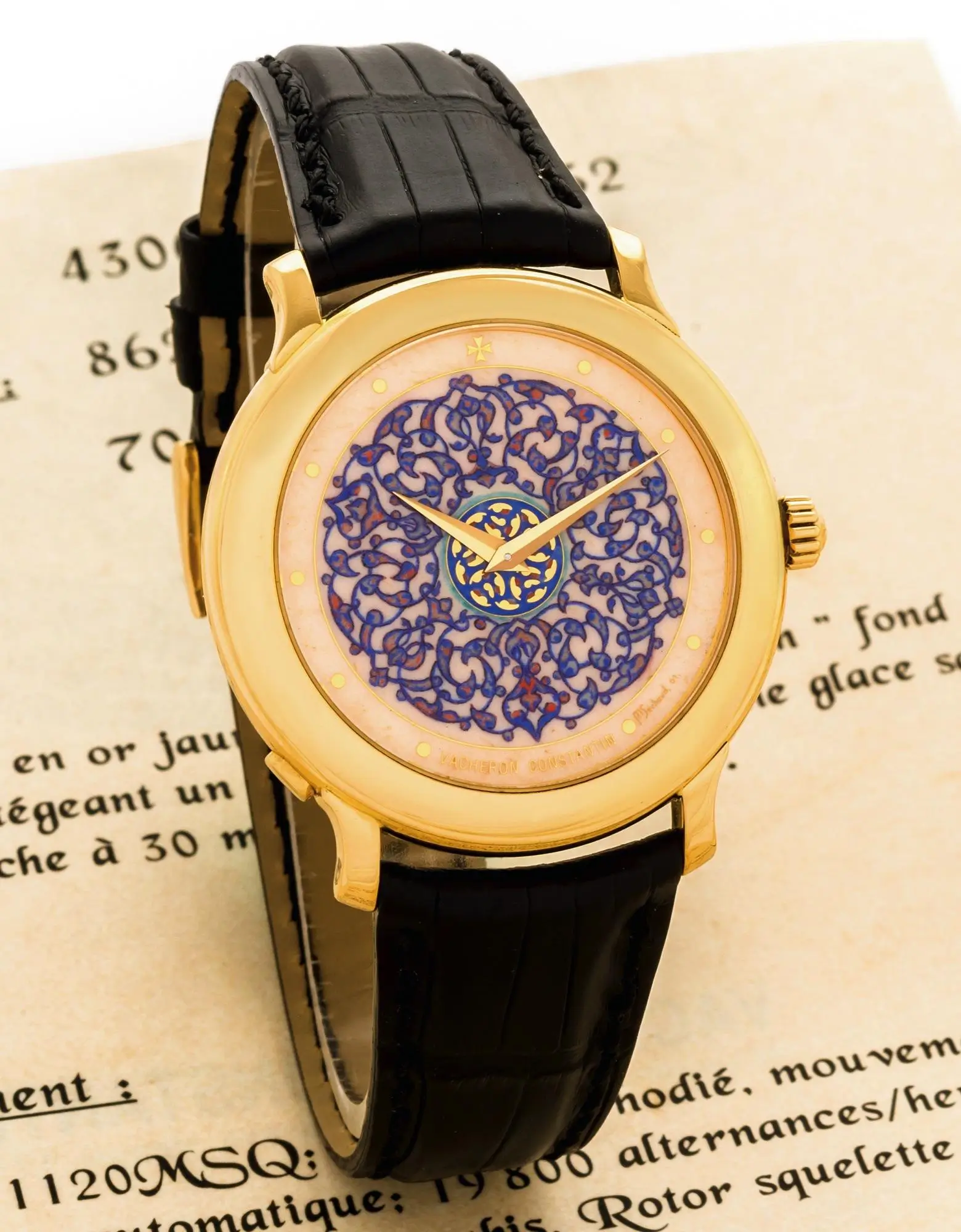 Gold wristwatch with black leather strap featuring an ornate blue enamel dial, showcasing intricate scrolling motifs and central medallion detail, set against a vintage document backdrop