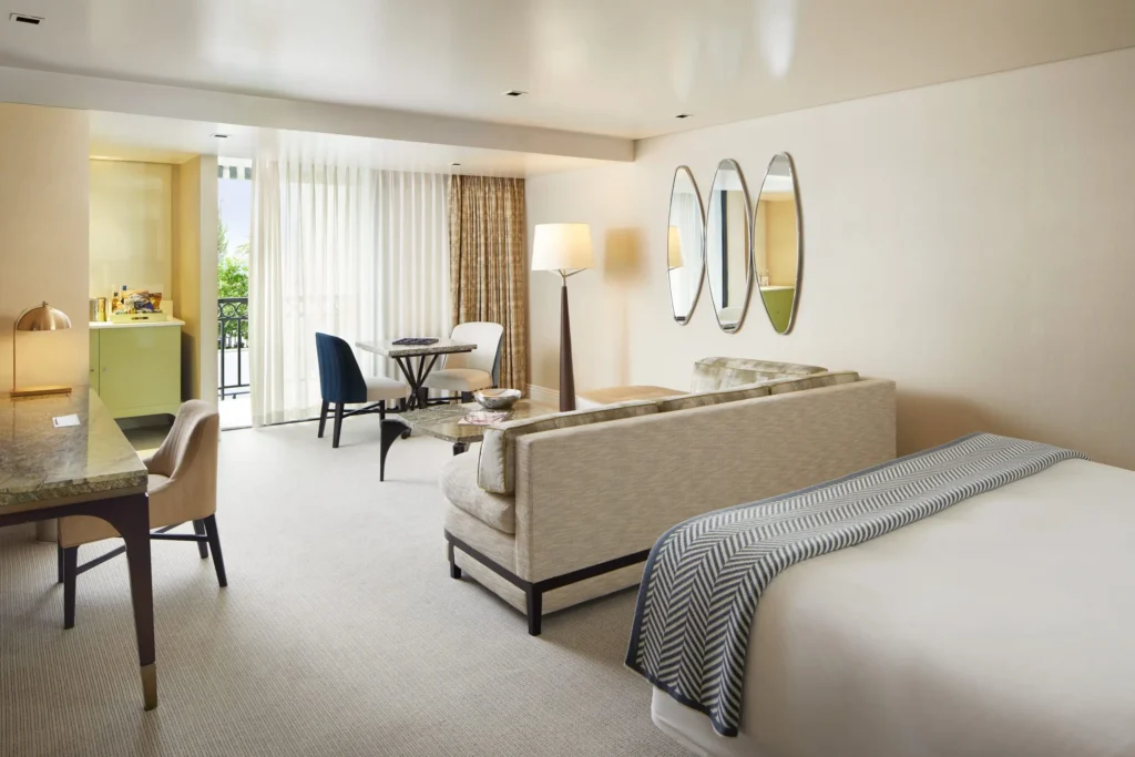 Spacious, softly lit hotel suite featuring a neutral-toned sitting area with a plush sofa, glass coffee table, and a bed in the foreground with a patterned throw. A small dining table with upholstered chairs sits near floor-to-ceiling sheer curtains leading to a balcony, while a marble desk, modern floor lamp, and trio of oval wall mirrors complete the refined, contemporary interior