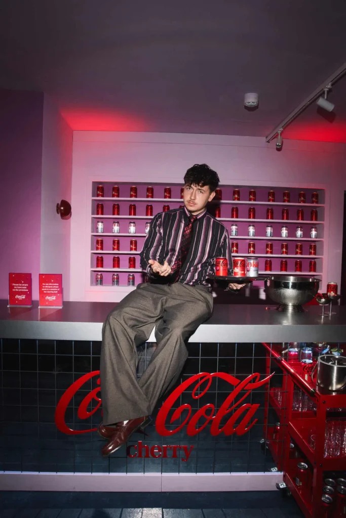 A stylized café interior captures a person seated casually on the countertop, framed by a wall of neatly arranged Coca-Cola Cherry cans that form a graphic, almost grid-like backdrop. Dressed in a striped button-up shirt with a coordinating tie and wide-leg trousers, they hold a tray of drinks while gesturing mid-conversation, giving the image a candid, editorial edge. The space is bathed in soft pink and red lighting, reinforcing the cherry-centric palette, while the glossy black-tiled counter—branded with a bold Coca-Cola Cherry script—anchors the composition. To the side, a metal bowl filled with cans and a red service trolley stocked with glassware add layers of function and styling. The overall scene blends hospitality and fashion, reading as both a lived-in moment and a carefully composed campaign visual