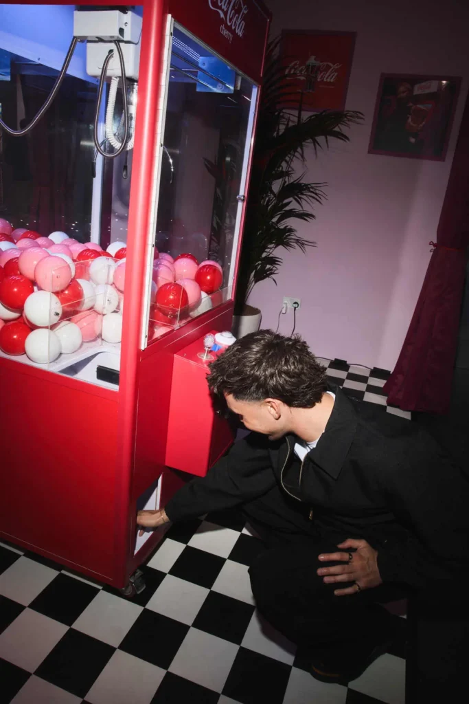 interaction as they reach toward the lower access panel—less about winning, more about curiosity and play. The machine itself is filled with glossy red, pink, and white spheres, echoing the Cherry Coke palette and turning a familiar arcade object into a branded centerpiece. The setting leans into contrast: a black-and-white checkered floor grounds the scene in retro diner nostalgia, while the surrounding lighting shifts between cool blue tones inside the machine and warm, saturated reds spilling across the room. A potted plant and framed wall art soften the otherwise graphic environment, adding a hint of lived-in texture. The subject’s posture—bent, focused, slightly amused—introduces a sense of spontaneity that breaks from the more posed, social imagery elsewhere. It feels like a behind-the-scenes beat in a larger experience, where interaction replaces performance. Overall, the image balances playful design with human curiosity: a tactile, almost mischievous engagement with the installation, reinforcing the idea that this space isn’t just to be seen—it’s meant to be explored.