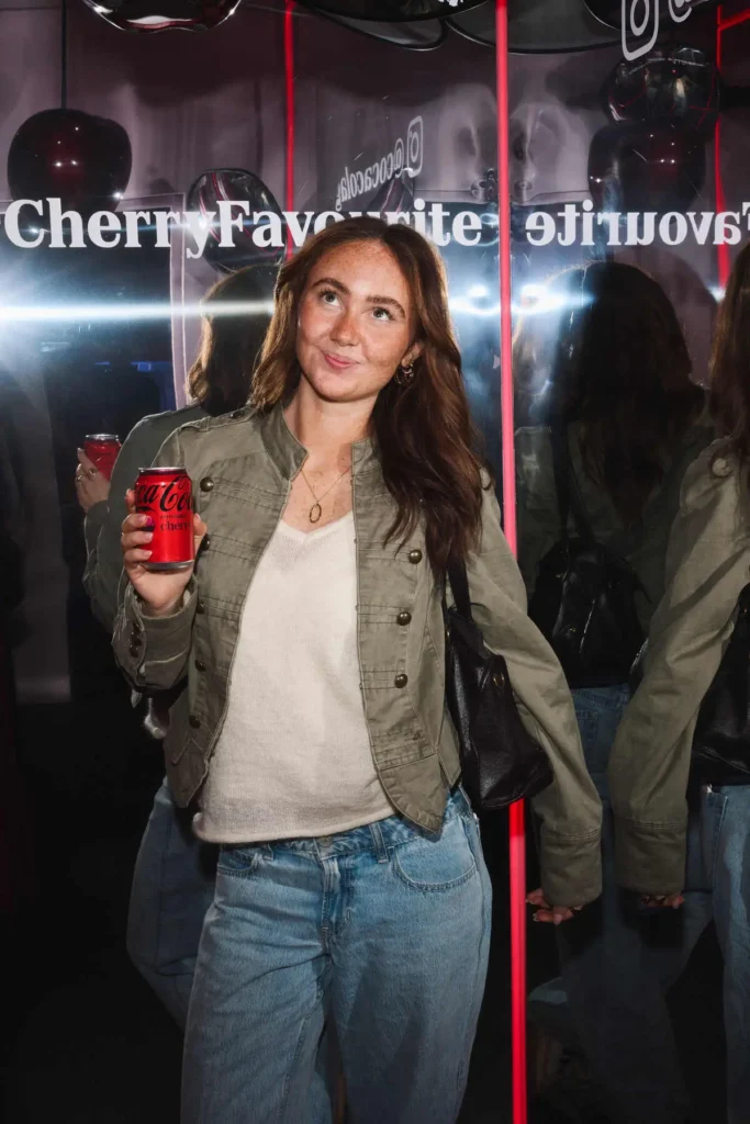 A dynamic, mirror-lined scene captures a person mid-step, holding a can of Coca-Cola Cherry as reflections multiply both their figure and the surrounding branding. Dressed casually in a light knit top, olive-toned jacket, and relaxed denim, they carry a shoulder bag while glancing upward with a playful, slightly posed expression. Behind them, bold white lettering—“Cherry Favourite”—appears reversed across the mirrored panels, intersected by vertical red light strips that echo the brand’s signature palette. The layered reflections and lighting create a sense of depth and motion, turning a simple moment into a visually immersive, campaign-ready composition