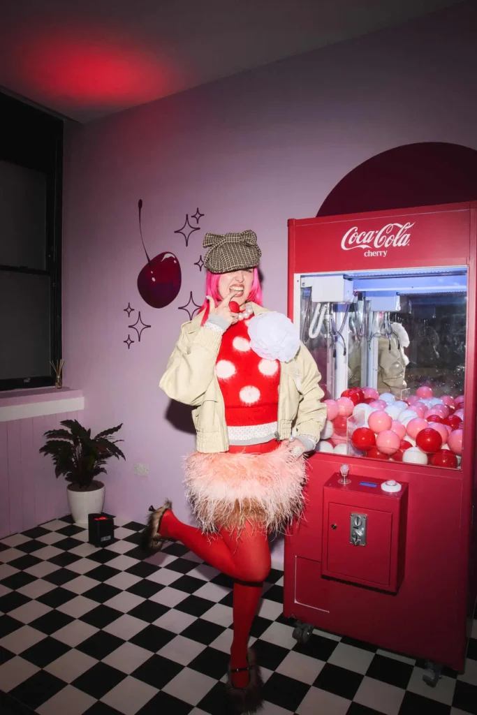 A high-energy, editorial-style scene captures a person posing playfully beside a Coca-Cola Cherry claw machine, fully immersed in a candy-colored, retro-inspired environment. With vivid pink hair tucked under a tweed cap, they strike a cheeky pose—balancing on one leg while biting into a small red ball that echoes the cherry theme. The outfit is deliberately maximalist: a bold red polka-dot sweater layered under a beige jacket with an oversized sculptural flower, paired with a feathery skirt and red tights. The setting reinforces the aesthetic—lavender walls with graphic cherry illustrations, a curved red backdrop framing the machine, and a black-and-white checkered floor grounding the look in diner nostalgia
