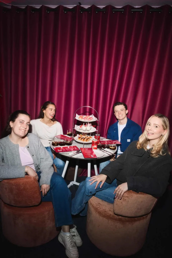 A cozy, lounge-style scene unfolds around a small circular table, where four people sit comfortably in plush, rounded chairs against a rich cherry-red curtain backdrop. The setting feels intimate and curated—somewhere between a café and a private tasting room—bathed in warm, flattering light that enhances the deep reds and soft neutrals throughout. At the center, a tiered stand presents an assortment of desserts and small bites—mini pastries, cakes, and neatly arranged treats—styled almost like an afternoon tea, but with a playful, branded twist. Around it, short glasses filled with dark soda and ice, alongside red Coca-Cola cans, anchor the experience firmly within the Cherry Coke theme. Menus in matching red are placed neatly on the table, reinforcing the cohesive visual identity. The group’s body language is relaxed and conversational—one leans back casually, another turns mid-laugh toward a friend—capturing a moment that feels both social and staged. Their outfits remain understated—denim, knits, and simple layers—allowing the saturated backdrop and curated table setting to take visual priority. The overall composition balances hospitality and branding: a stylized café moment that feels shareable, inviting, and quietly indulgent, where the product integrates seamlessly into a broader lifestyle vignette