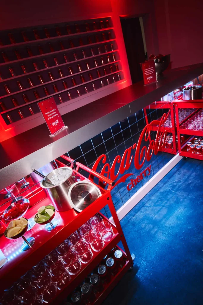 A sharply angled, almost cinematic view of the service area reveals the operational core of the Cherry Coke café—where styling meets function. The composition leans diagonally, giving the space a dynamic, editorial energy as the eye moves from foreground to background in a clean, continuous sweep. In the foreground, a vivid red service cart is meticulously arranged: rows of empty glasses line the lower shelves, while the top tier holds a metal ice bucket, sliced citrus, and a bowl of glossy cherries—ingredients that suggest both garnish and ritual. Beneath, neatly stacked Coca-Cola Cherry cans reinforce the product’s presence without overwhelming the frame. The bar counter stretches across the middle ground, sleek and metallic, with small red signage placed deliberately along its surface. Below it, a tiled front panel features the iconic Coca-Cola script in bold red, glowing subtly under the ambient lighting. Behind, a wall of evenly spaced cans forms a grid-like display—part inventory, part visual installation—lit by saturated red light that washes the entire space in a cohesive hue. The deep blue floor contrasts sharply with the red-dominant palette, grounding the scene and adding depth. There are no people here, which shifts the focus to environment and preparation. It feels like a pause between moments—everything set, waiting for interaction. Overall, the image captures the infrastructure of the experience: precise, branded, and visually striking, where every surface—from cart to counter to wall—works in sync to extend the Cherry Coke identity into a fully immersive spatial language