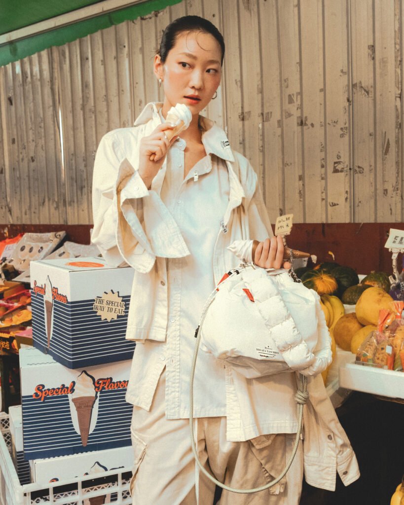 A young woman stands in a market setting, wearing an oversized off-white button-up shirt layered over loose, matching trousers. Her hair is pulled back neatly, and she holds a soft-serve ice cream close to her lips, mid-bite, with a calm, slightly contemplative expression. In her other hand, she carries a padded, quilted white handbag with rope detailing, suggesting a fashion-forward, utilitarian design. Behind her, stacked boxes labeled “Special Flavor” and a display of fresh produce—melons, bananas, and other fruits—create a contrast between everyday market life and elevated, minimalist styling. The lighting is warm and slightly diffused, giving the scene a candid, editorial feel