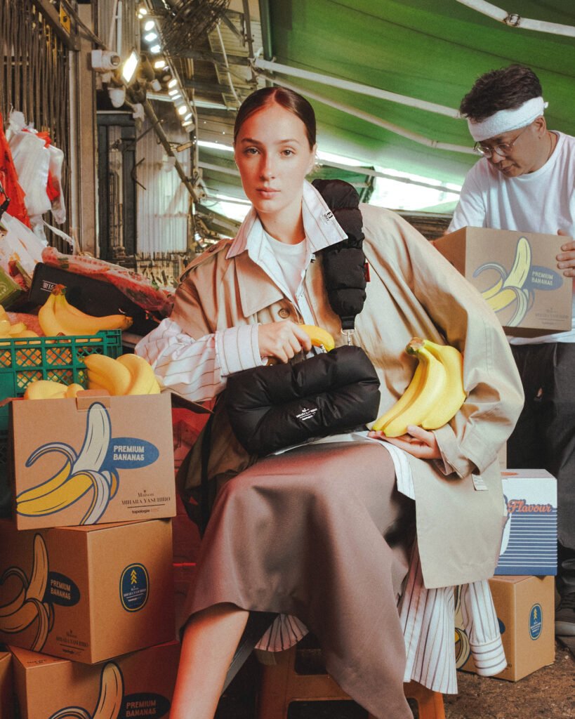 A woman stands in a market workspace surrounded by stacked green produce boxes, wearing a beige trench-style coat layered over a striped shirt and neutral-toned skirt. Her hair is neatly tied back, and she looks downward with a focused, composed expression. In one hand, she holds a black, padded handbag with a soft, quilted structure and a thick, rounded handle, while a rope-style strap with orange accents crosses her body. A piece of fruit is tucked playfully into the bag’s opening, blending utility with styling. Beside her, a pallet jack adds an industrial element to the scene, reinforcing the working-market environment. The lighting is cool and slightly diffused, highlighting the contrast between the bag’s matte texture and the structured surroundings