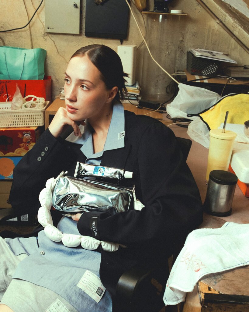 A woman sits in a cluttered, backroom workspace, dressed in an oversized black blazer layered over a light blue collared dress. Her hair is pulled back neatly, and she rests her chin on her hand while gazing off to the side with a thoughtful, slightly distant expression. Cradled in her lap is a compact, metallic silver bag with a glossy, reflective finish, paired with a soft, padded white strap that loops around it. A tube—resembling toothpaste—rests across the top of the bag, adding a subtle, unconventional detail. The surrounding environment includes scattered objects like paper bags, containers, cables, and a drink cup, creating a lived-in, utilitarian atmosphere. Warm, diffused lighting softens the scene, balancing the contrast between the polished accessories and the raw, everyday setting