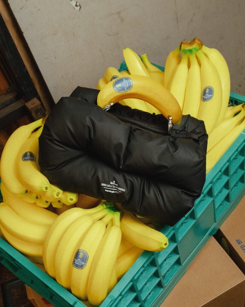 A black, padded handbag with a soft, pillowy construction rests inside a plastic crate filled with bright yellow bananas. The bag’s rounded, cushioned form contrasts with the smooth, curved shapes of the fruit surrounding it, while a banana is hooked through the bag’s opening like a playful, improvised handle. Subtle branding is printed on the front of the bag, maintaining a minimalist aesthetic. The crate’s teal color and the vivid yellow of the bananas create a bold, saturated backdrop, emphasizing the bag’s matte black finish. The composition blends everyday produce with stylized presentation, giving the scene a conceptual, editorial quality