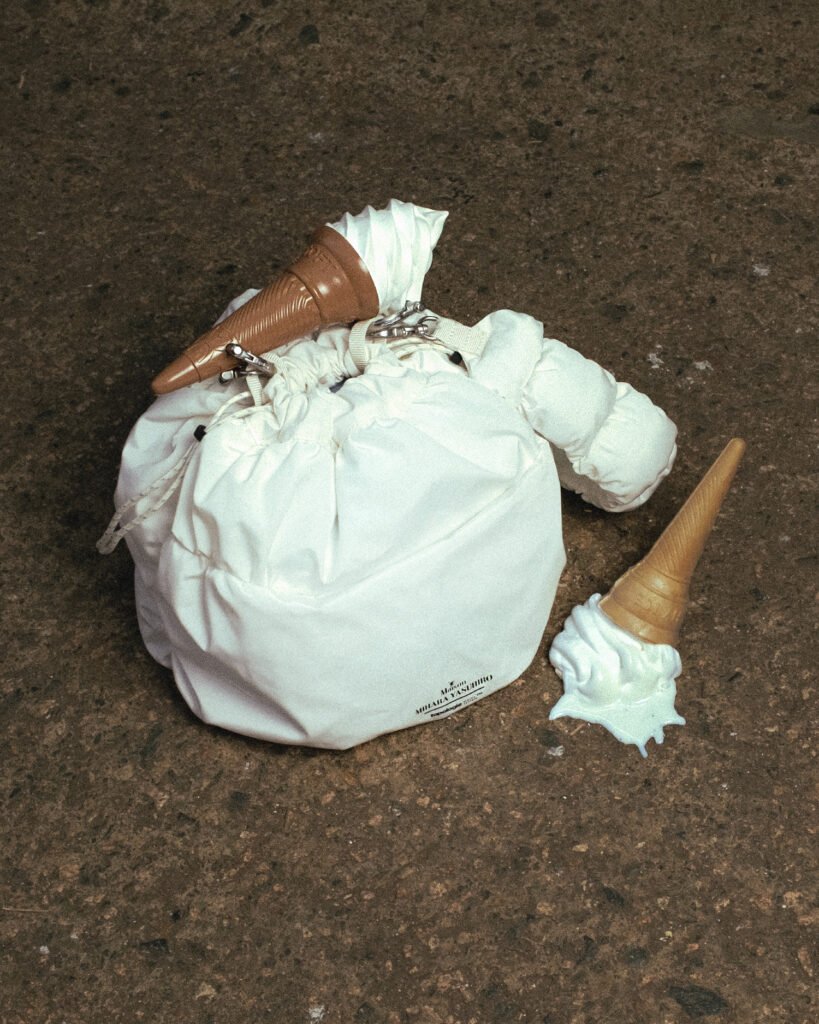 A white, padded drawstring bag with a soft, pillowy structure rests on a rough concrete floor, its fabric gathered at the top with visible cords and metal clips. A thick, cushioned strap loops around the bag, reinforcing its sculptural, utilitarian design. Resting on top is an intact soft-serve ice cream cone, while another lies beside it on the ground, tipped over with the ice cream partially melted and spilling outward. The contrast between the pristine, minimal bag and the messy, melting cones creates a striking, conceptual composition. Warm, slightly muted lighting emphasizes the textures—the smooth swirl of the ice cream, the matte softness of the bag, and the gritty surface beneath