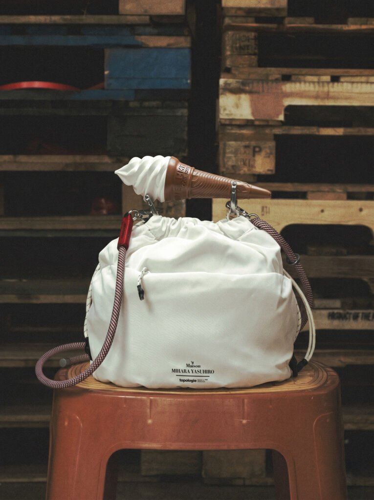 A white, padded drawstring bag with a soft, gathered silhouette sits centered on a plastic stool, its surface smooth and lightly structured with visible seams and a front pocket. A rope-style strap with red accents loops from either side, adding a technical, utilitarian detail to the otherwise minimal design. Balanced across the top opening is a soft-serve ice cream cone, placed horizontally and secured by the bag’s metal clips, introducing a playful, unexpected element. The background features stacked wooden pallets and an industrial storage setting, contrasting the bag’s clean, refined appearance with a raw, textured environment. Warm, muted lighting enhances the softness of the bag’s material while subtly highlighting the glossy swirl of the ice cream and the worn surfaces behind it