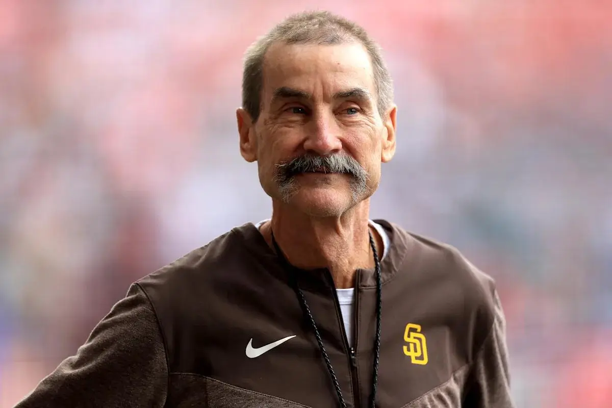 A close-up portrait of a middle-aged Peter Seidler with short graying hair and a thick mustache, wearing a brown San Diego Padres zip-up jacket with a Nike logo and team emblem, standing against a softly blurred stadium background