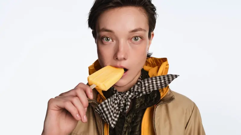 Close-up portrait of a model in Moncler summer layers, wearing a beige lightweight jacket over a vivid yellow hooded layer with a gingham scarf, holding a yellow ice pop, reinforcing the campaign’s playful, warm-weather mood