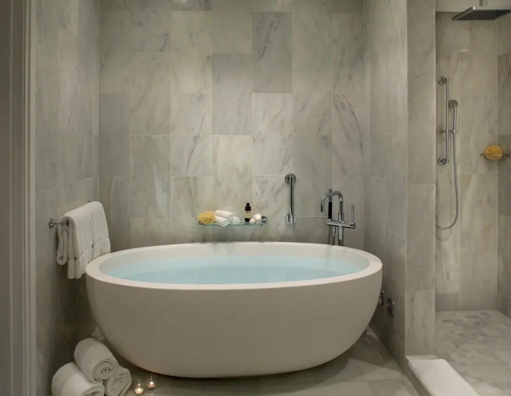 Minimalist spa-style bathroom featuring a freestanding oval soaking tub filled with water, set against large-format marble tile walls. A sleek chrome floor-mounted faucet and a glass shelf with neatly arranged bath essentials sit above the tub, while rolled white towels and small candles add a calming touch. To the side, a walk-in rainfall shower with matching marble finishes completes the serene, high-end setting