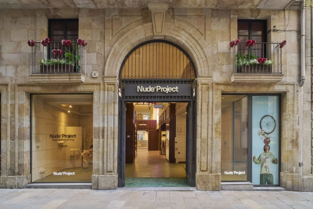 A street-level view of a historic stone building housing a Nude Project retail store, framed by an arched entrance with black signage above. Large display windows on either side reveal minimalist interiors, including a visible installation and a poster-style visual featuring a figure with a bicycle wheel. The left window reads “Nude Project New store opening 25.04.26.” Inside, warm lighting illuminates a long, corridor-like layout with wood finishes. Small balconies above the storefront are decorated with plants and sculptural red elements, adding a play contrast to the aged stone façade