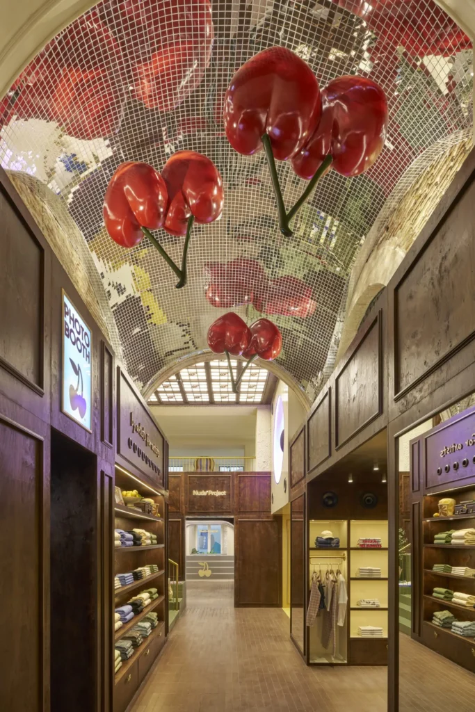 An interior view of a boutique retail space with warm wood-paneled walls and neatly arranged clothing displays on both sides. Above, a curved mirrored ceiling is tiled in a mosaic pattern, reflecting oversized glossy red cherry sculptures mounted upside down, creating a surreal, immersive canopy. Soft lighting highlights folded garments, hanging pieces, and built-in shelving, while the corridor leads toward a central back area marked with Nude Project branding, blending playful design with a refined, gallery-like retail environment