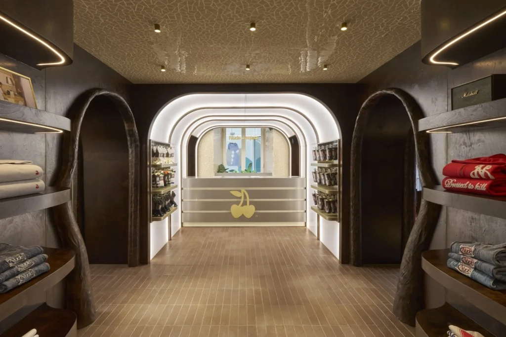A symmetrical view inside a contemporary boutique featuring a softly illuminated, arched passage leading to a central counter adorned with a cherry logo. Dark wood-paneled walls frame curved alcoves on both sides, where neatly folded clothing and accessories are displayed on built-in shelves. Warm recessed lighting and sculptural ceiling patterns create a refined, immersive atmosphere, while the clean lines and glowing archway guide the eye toward the back of the store