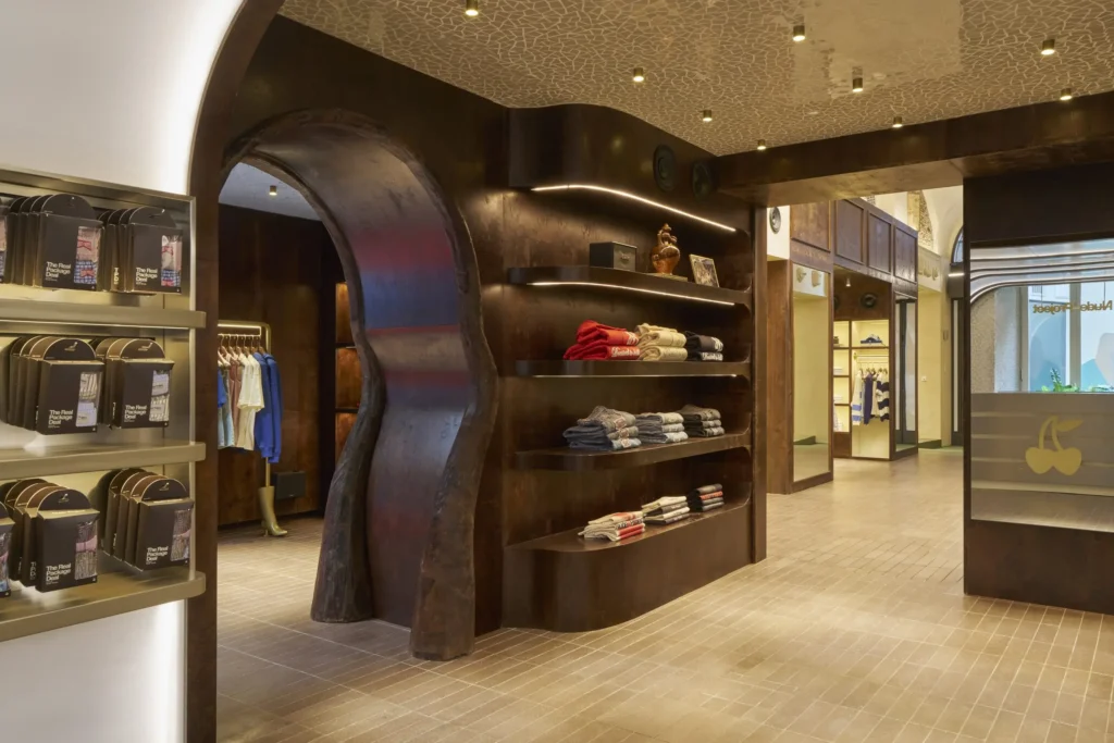An interior view of a boutique space featuring sculptural, dark wood architecture with a curved, organic doorway leading into a clothing area. Built-in shelves display neatly folded garments in neutral and bold tones, highlighted by warm integrated lighting. To the left, a lit shelving unit presents boxed accessories, while the open layout extends toward additional rooms and a central counter marked by a cherry logo. Textured flooring and a patterned ceiling with recessed lights enhance the refined, immersive retail environment
