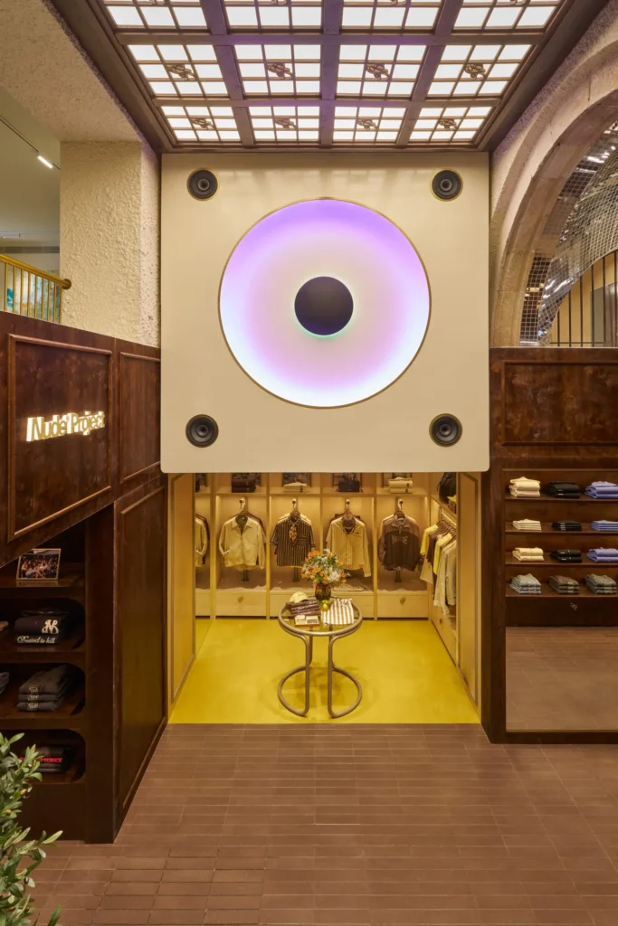 A boutique interior centered on a large square installation featuring a glowing circular gradient light resembling an abstract eye, mounted above a display niche. Beneath it, a small round table with books and a floral arrangement sits on a bright yellow floor, surrounded by neatly hung garments. Rich wood-paneled walls with built-in shelving frame the space, while a grid-patterned skylight above casts soft, diffused light, blending art installation with curated retail design