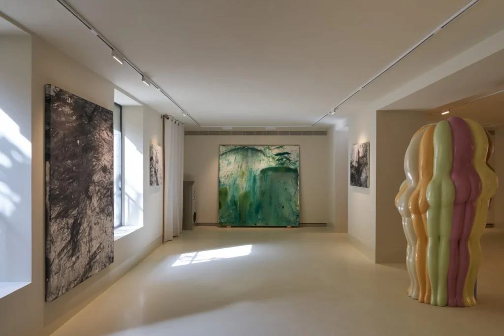 A minimalist gallery-like interior with white walls and soft natural light streaming through a side window. Large abstract paintings hang along the walls, including a prominent green-toned canvas at the far end. In the foreground to the right stands a glossy, sculptural installation composed of rounded, elongated forms in pastel colors, resembling clustered human silhouettes. Track lighting and clean architectural lines create a calm, contemporary exhibition atmosphere