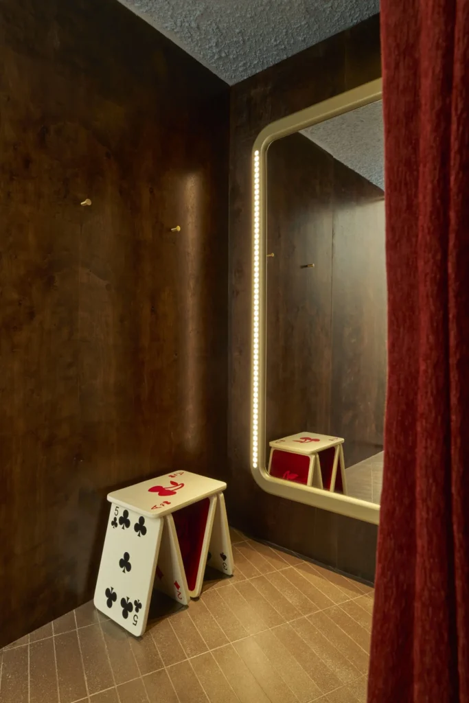 A fitting room corner with dark wood-paneled walls and a large, softly illuminated mirror framed by rounded edges and vertical lights. In front sits a sculptural stool designed like oversized playing cards, featuring club and heart symbols with a red interior. A heavy red curtain partially frames the scene, while warm lighting and textured flooring create an intimate, stylized dressing area