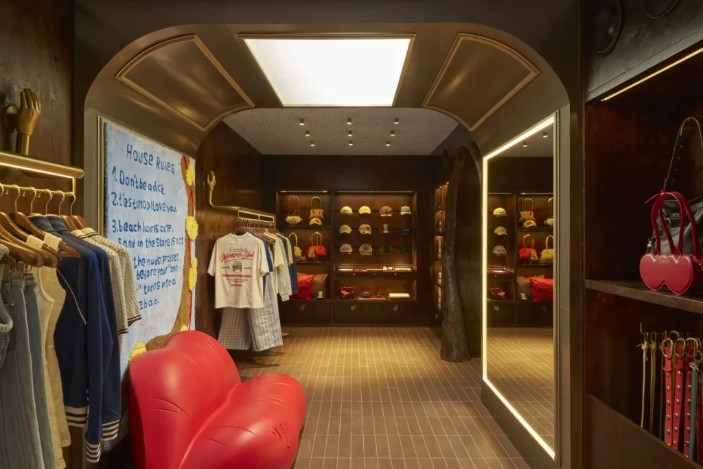 A boutique interior with rich wood-paneled walls and warm lighting, featuring a curved, metallic archway leading into a display area. On the left, clothing hangs neatly beside a large textile artwork with handwritten-style text, while a sculptural red chair shaped like a heart sits in the foreground. On the right, a tall illuminated mirror reflects shelves of hats and accessories, and a display of red leather goods adds contrast. The space blends playful design elements with a refined retail layout