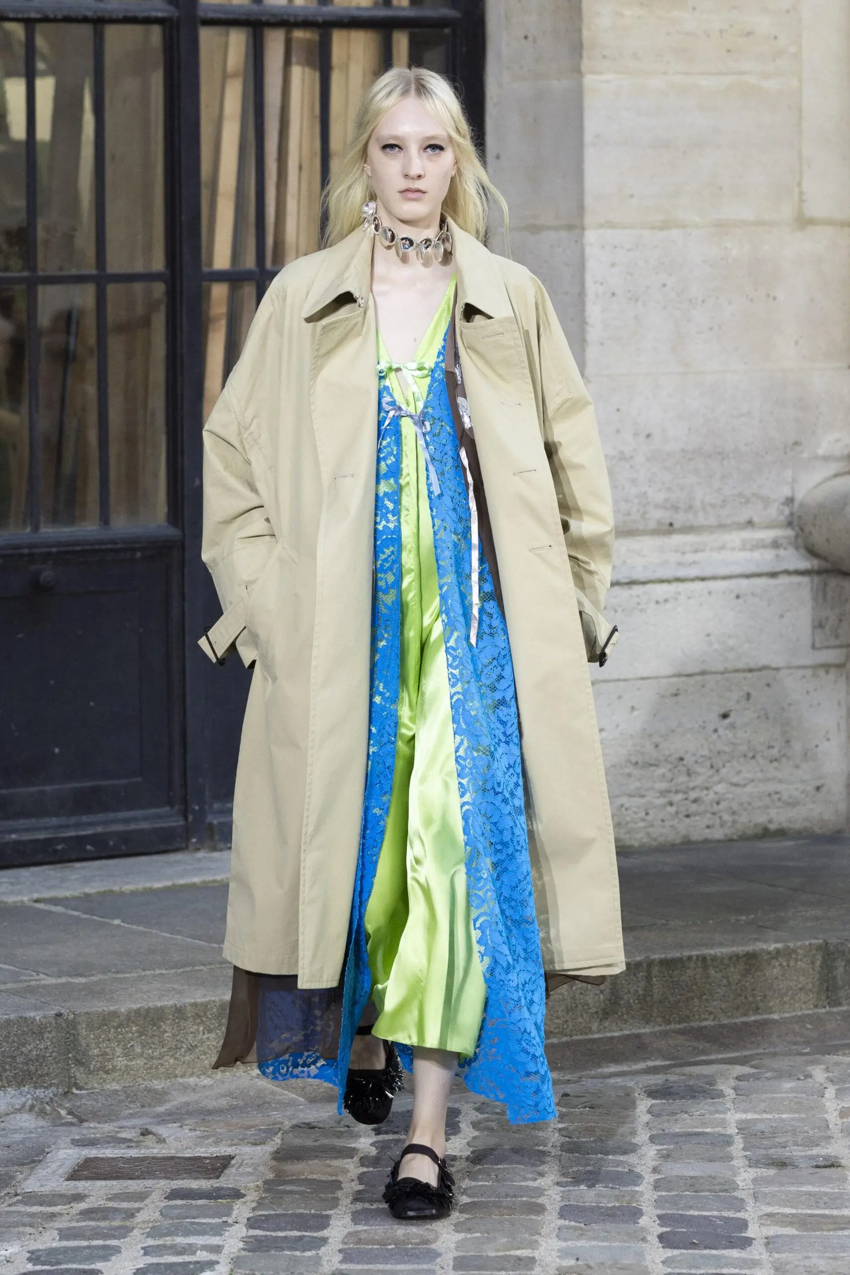 Full-body runway-style image of a model walking along a cobblestone street, framed by a stone façade and dark window panels. She wears an oversized beige trench coat with a relaxed, fluid silhouette, left open to reveal layered textures and bold color beneath. Underneath, a vivid lime-green satin slip dress catches the light with a soft sheen, overlaid with a semi-sheer blue lace layer that adds depth and contrast. The neckline features delicate ribbon ties, echoing a romantic, lingerie-inspired influence. The look is accessorized with a chunky statement choker composed of large, reflective elements, adding a sculptural, almost metallic accent at the collar. Her footwear consists of black flat shoes with subtle embellishment, keeping the outfit grounded. Hands tucked casually into the coat pockets, the model’s styling feels effortless yet intentional—balancing utilitarian outerwear with vibrant, feminine layering. The overall composition merges softness and structure, set against an understated urban backdrop