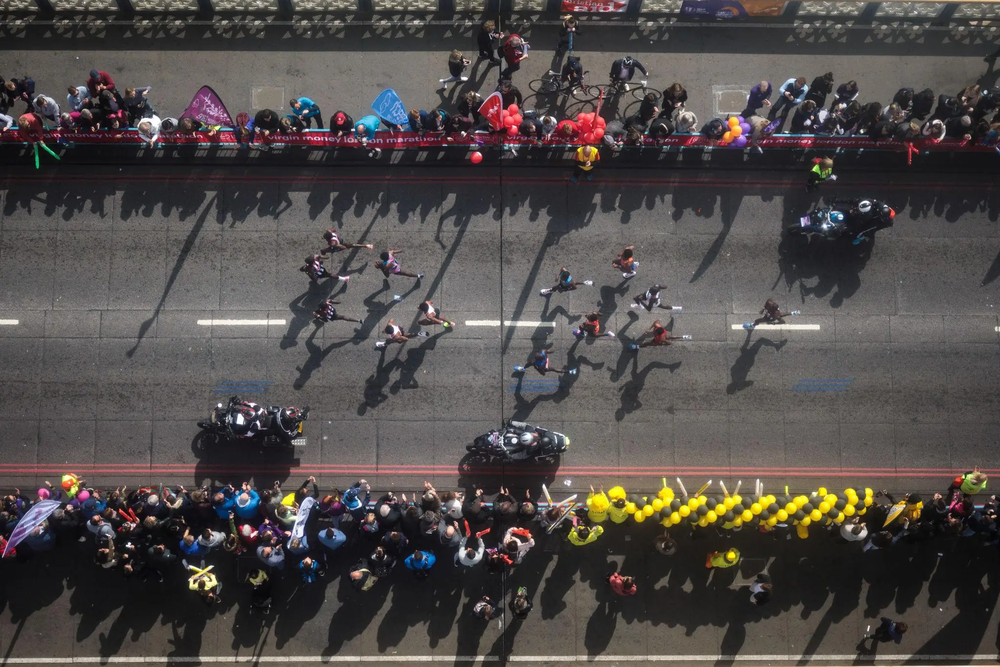 Aerial view of a city marathon in progress, with runners spread across a wide road flanked by dense crowds on both sides. Spectators lean over barriers holding balloons and banners, while long shadows stretch across the pavement in bright sunlight. Several motorcycles escort the runners, and the scene captures a dynamic contrast between motion on the course and the stationary audience above and below