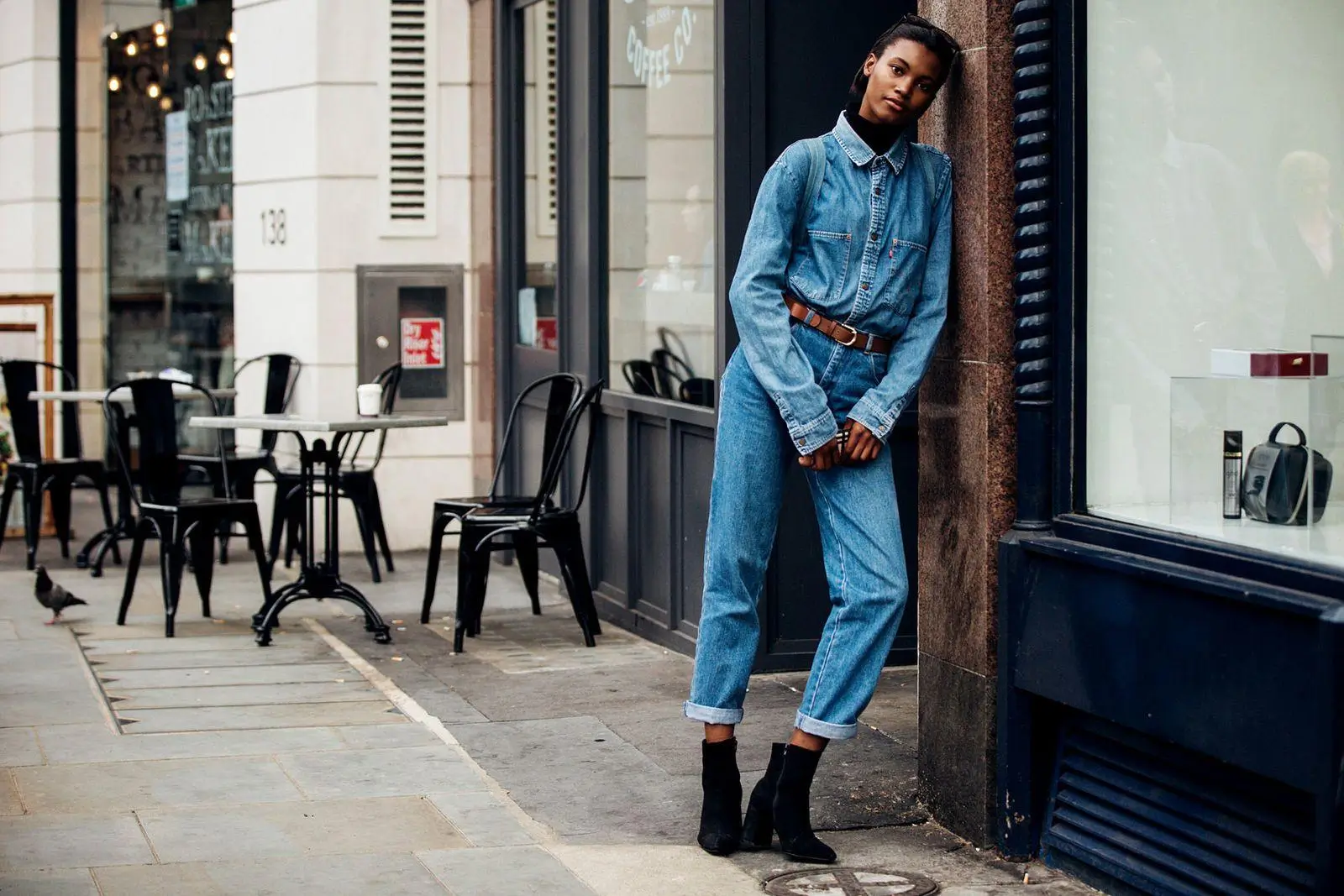 A model stands on a city sidewalk leaning against a storefront, wearing a full denim outfit with a belted shirt and cuffed jeans paired with black ankle boots; behind her, café tables and chairs line the street, adding a casual urban atmosphere