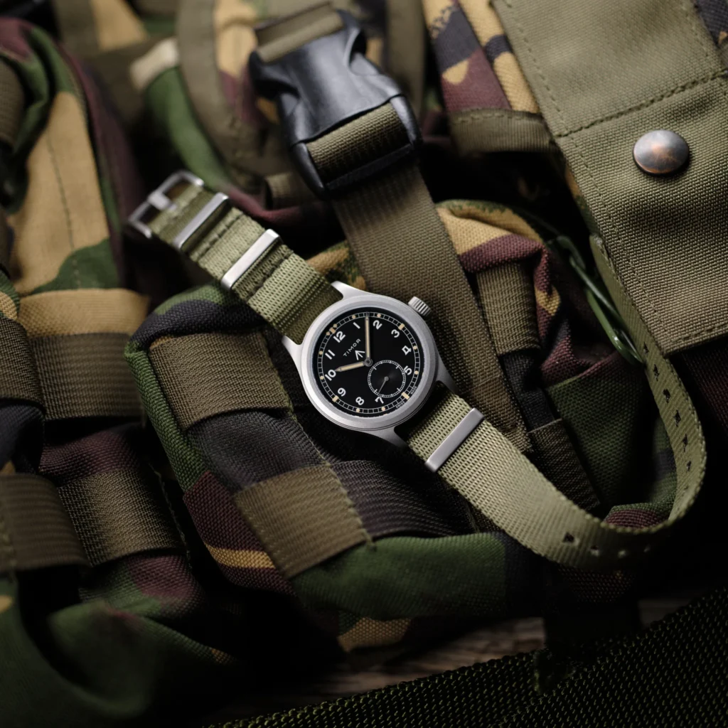 A close-up of a rugged field watch with a black dial and olive green fabric strap, resting among camouflage-patterned gear and nylon webbing, evoking a military-inspired, utilitarian aesthetic